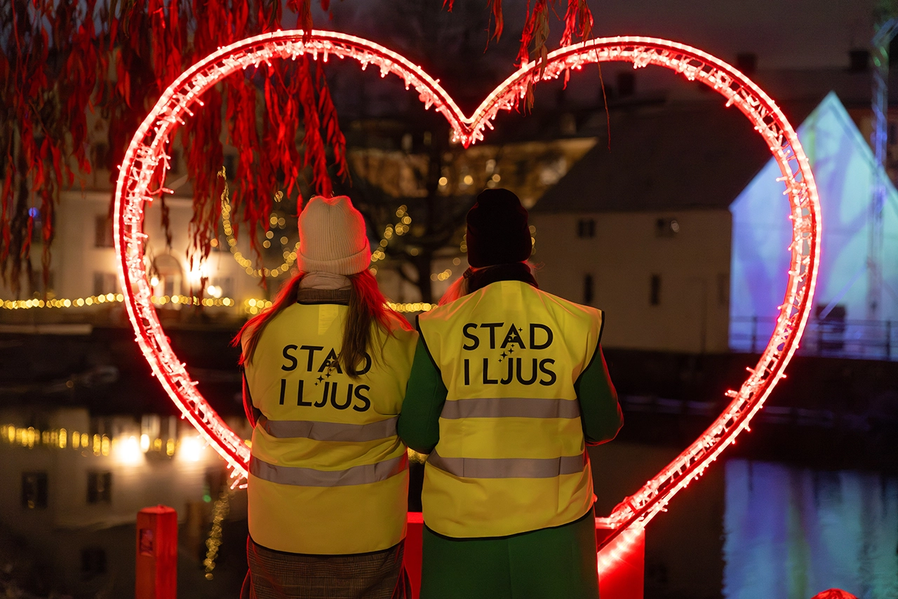 Två personer står framför ett rött lysande hjärta med reflexvästar med texten "Stad i Ljus".